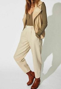 Beige kožená vesta přes žebrované olivově zelené tričko, zkombinovaná s krémovými širokými kalhotami a hnědými kotníkovými botami.