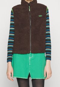 Brauner Fleece-Weste mit hohem Kragen und grünem Reißverschluss, getragen über einem gestreiften Langarmshirt und einem grünen Rock. Auffälliges Levi's-Logo auf der Brust.