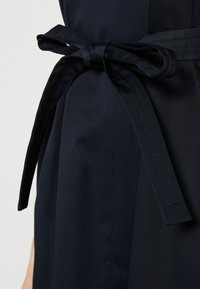 Gros plan sur une ceinture en tissu noir nouée en un nœud autour de la taille d'un vêtement sombre sur un fond uni.