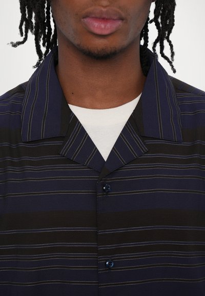 Chemise à rayures noir marine et noires, boutonnière à l'avant avec des boutons bleus, col court et tissu texturé. Superposée sur un T-shirt blanc.