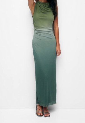 Femme portant une robe maxi sans manches ajustée vert ombré avec des côtés froncés et des talons argentés à fines lanières sur un fond blanc uni.