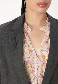 Grå rutete blazer lagd over en bluse med blomsterprint i nyanser av rosa, oransje og grønt. Svart sikkerhetsnål som aksent på klaffen.