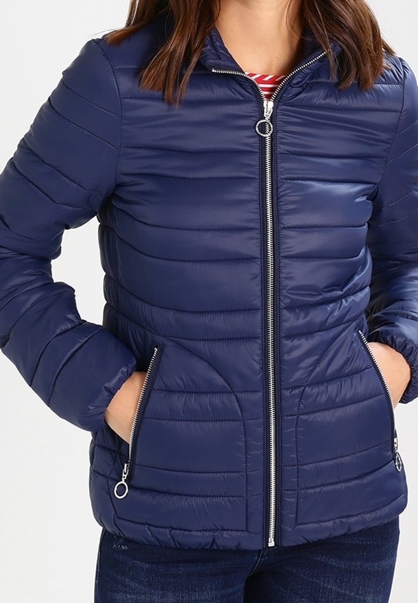 Femme portant une veste matelassée bleu marine avec des poches zippées argentées et une fermeture éclair frontale, les mains dans les poches, par-dessus une chemise rayée rouge et blanche.