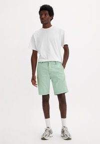 T-shirt bianca in cotone, pantaloni corti verde chiaro con una vestibilità rilassata e sneakers grigie. Il modello sta in piedi con le mani in tasca su uno sfondo semplice.