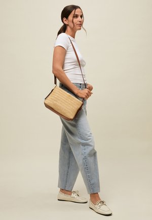 Mujer con cabello oscuro recogido en una coleta, vistiendo una camiseta blanca, vaqueros azul claro, mocasines crema y llevando un bolso cruzado tejido color cuero claro.