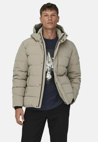 Beige puffer-takki, jossa on huppu, tyyliltään tikattu, edessä napitus ja mustat yksityiskohdat, päällä tummansininen graafinen huppari.