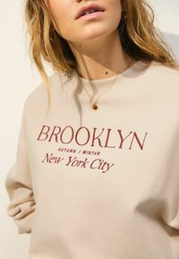 Beige sweatshirt met rode geborduurde tekst: "BROOKLYN AUTUMN / WINTER New York City." Ronde halslijn, zachte textuur, losvallende pasvorm.