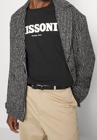 Svart och vit randig stickad kavaj med snedskuren krage, bärs över en svart grafisk T-shirt, tillsammans med beige byxor och ett svart bälte.