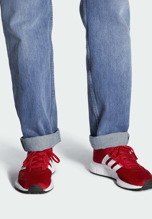Czerwone buty sportowe z białymi akcentami, tekstylnym wierzchem i amortyzowaną podeszwą. Noszone z podwiniętymi niebieskimi dżinsami.