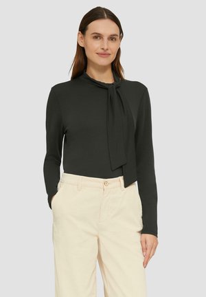 Vrouw met lang bruin haar, gekleed in een donkere, lang-sleeve top met een gestrikt nekomslag en lichtbeige hoge taille broek met haar handen in de zakken.