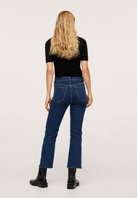 Mujer rubia vestida con una blusa negra de manga corta, jeans acampanados cortos de color azul oscuro y botas negras hasta el tobillo, de pie con las manos a los lados.