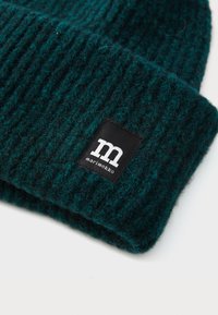 Berretto in lana a coste di colore verde scuro con risvolto, dotato di un'etichetta nera con la scritta bianca "m" e "marimekko" su sfondo bianco.