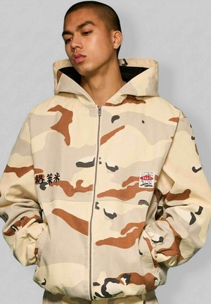 Osoba ubrana w beżowo-brązową kurtkę moro z kapturem i zamkiem błyskawicznym, z rękami w kieszeniach, patrząca w dół na jednolitym tle.