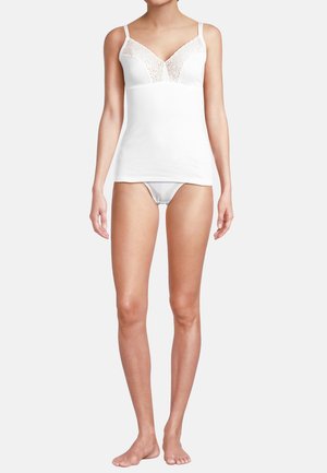 Nainen valkoisessa pitsireunaisessa topissa ja siihen sopivissa bikinialusvaatteissa, seisomassa paljain jaloin vaaleaa taustaa vasten.