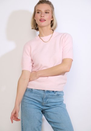 Frau trägt einen hellrosa kurzärmeligen Pullover, eine goldene Kettenhalskette und hellblaue High-Waist-Jeans vor einem weißen Hintergrund.