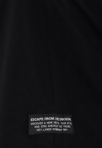 Tela negra con una etiqueta rectangular cosida que dice: "ESCAPA DE LO DESCONOCIDO, descubre un nuevo si* para tu estilo, mantente a la vanguardia de las tendencias, Key Largo est 1994."