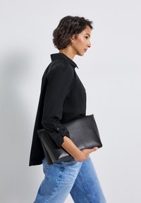 Donna con capelli corti e ricci che indossa una maglietta nera e jeans blu, mentre tiene una grande borsa a mano nera e cammina verso destra.