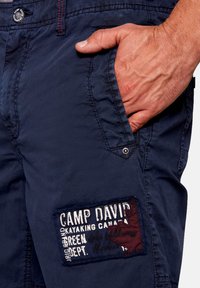 Sötétkék nadrág, struktúrált anyagból, oldalsó zsebbe és egy téglalap alakú címkébe, amelyen a "CAMP DAVID" márkaneve és kontrasztos színek találhatók.