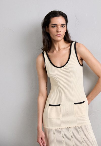 Top sin mangas de punto en color crema con textura acanalada, ribete negro alrededor del escote y dos bolsillos con detalles negros.