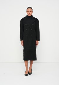 Manteau noir croisé avec des revers surdimensionnés, manches longues et longueur midi droite. Fabriqué à partir d'un tissu texturé avec de gros boutons.