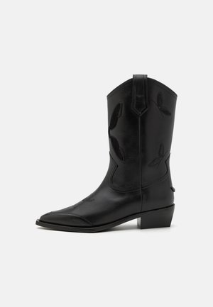 Botte de cowboy en cuir noir avec un bout pointu, des accents brodés et un talon bas. Présente des coutures verticales et des languettes de tirage sur les côtés.