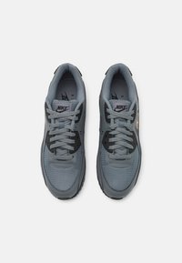 Grå Nike-sneakers med övre del av nät, svarta accenter, runda snören och gummisula, med en diskret logotyp på tungan och sidan.