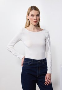 Weißes Langarmoberteil mit einem runden Halsausschnitt, kombiniert mit dunkelblauen High-Waist-Jeans. Glattes Material und figurbetontes Design.