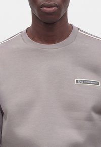 Άνδρας που φοράει ανοιχτό γκρι φούτερ Emporio Armani EA7 με λογότυπο στο στήθος και ριγέ σχεδιασμό στους ώμους, σε απλό φόντο.