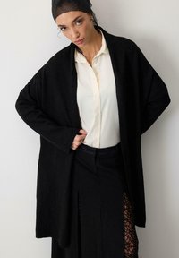 Melna pārslēgtā džemperis, izgatavots no teksturētas auduma, ar brīvu piegriezumu un platajām piedurknēm. Valkāts virs baltas blūzes un melnas svārku ar mežģīņu detaļām.