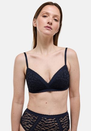 Femme portant un ensemble assorti de bralette en dentelle noire et culotte taille haute en dentelle, debout devant un fond clair uni.