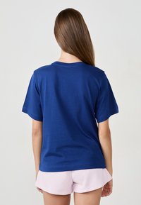 Femme aux longs cheveux bruns portant un T-shirt bleu uni à manches courtes et un short rose clair, de dos devant un fond uni.