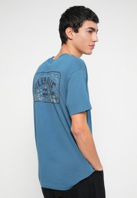 Blå bomulds-T-shirt med et grafisk design på bagsiden med træer og tropiske elementer. Korte ærmer og rund hals.