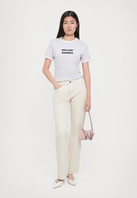 Tricou alb cu text negru "BRILLANT MADNESS", pantaloni foarte largi de culoare deschisă și o poșetă mică roz. Modelul stă cu încredere.