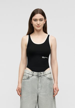 Noor naine pikkade pruunide juustega kannab musta varrukateta kombinesooni valge äärisega ja helehallide laia säärega teksapükse, seisab ühetaolise tausta ees.