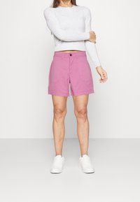 Shorts en coton rose avec fermeture à bouton, poches avant et coupe décontractée, assortis à un haut gris à manches longues et des baskets blanches.