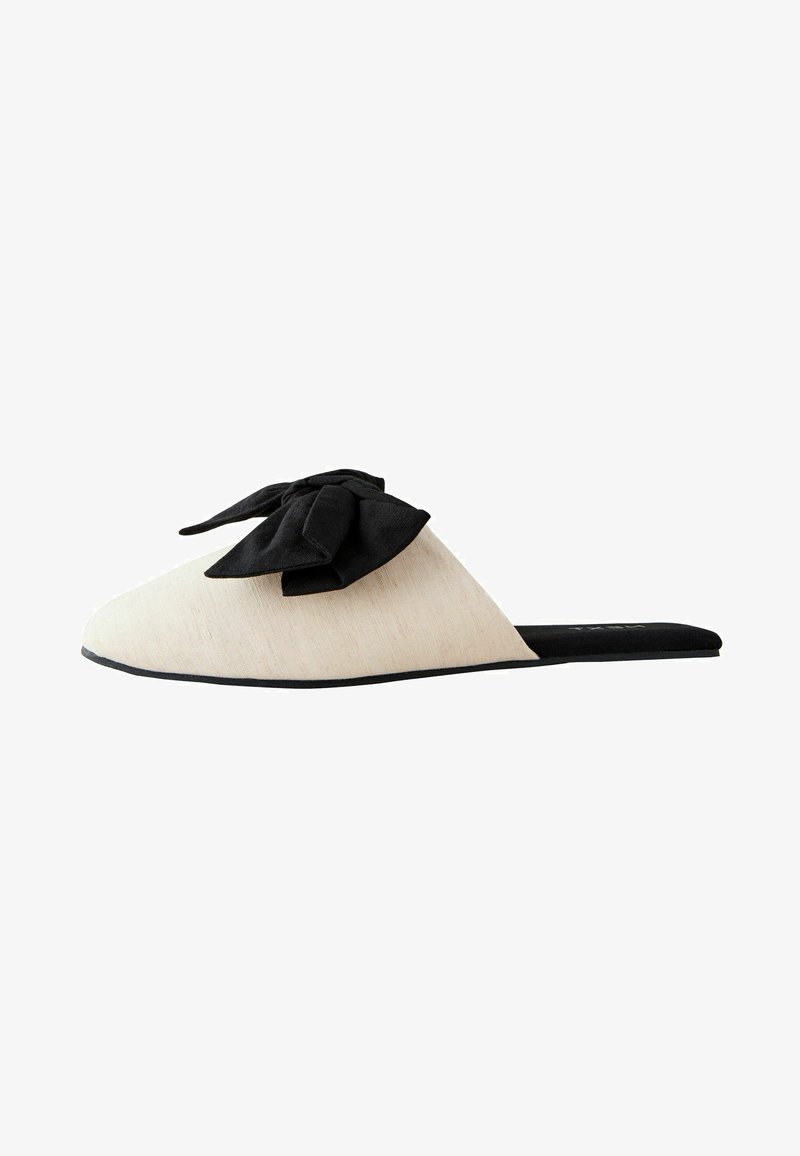 Pantofole beige senza lacci con un fiocco in tessuto nero sulla parte superiore. Presentano una superficie texturizzata e una suola nera piatta.