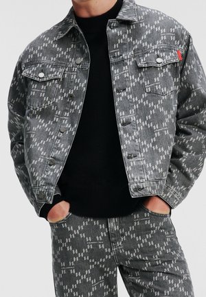 Mand iført matchende grå denimjakke og bukser med hvidt geometrisk mønster og lille tekst, hænderne i lommerne, sort sweater under.