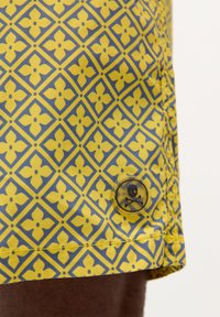 Pantaloni de baie galbeni cu un model floral albastru, având un buton transparent cu detaliu de craniu și oase încrucișate pe tiv.