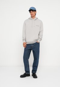 Sweatshirt gris clair avec une poche avant et une capuche à cordon, associé à un jean bleu ample et des chaussures noires. Casquette bleue avec un logo.
