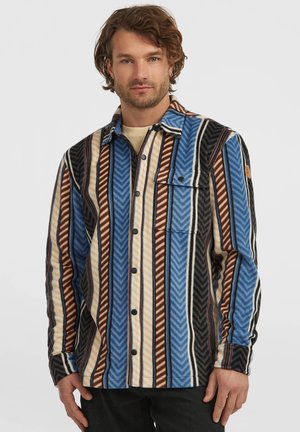 Man wearing a blue, brown, beige, and black vertically striped button-up shirt with a chest pocket, standing against a white background.