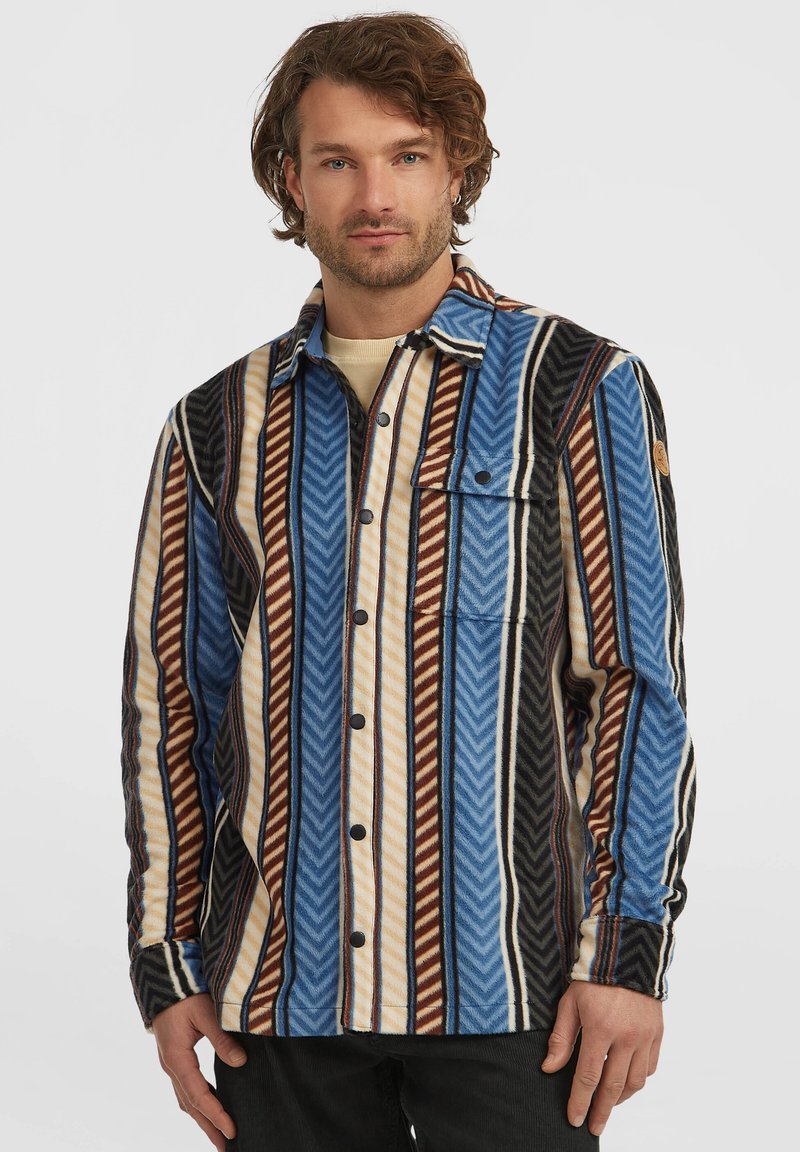 Homme portant une chemise à boutons à rayures verticales bleues, brunes, beiges et noires avec une poche sur la poitrine, se tenant devant un fond blanc.