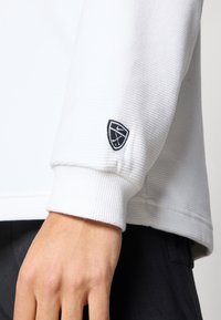 Λευκό ριμπ πλεκτό φούτερ μακρυμάνικο με στρογγυλή λαιμόκοψη και μαύρο κεντημένο λογότυπο στον δεξί καρπό. Μαλακή υφή και άνετη εφαρμογή.