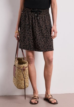 Femme portant une jupe imprimé léopard, un top noir, un bracelet doré, des sandales noires et dorées, tenant un sac cabas en paille tissé avec des anses en cuir.