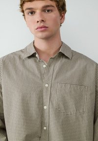 Braun- und cremefarbendes kariertes Hemd mit langen Ärmeln, Button-down-Kragen, Brusttasche und weißen Knöpfen auf einem untexturierten Hintergrund.