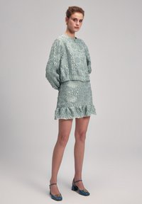 Conjunto de dos piezas de tela texturizada verde claro que incluye una blusa bordada de mangas largas y una falda acampanada con un dobladillo con volantes. Tacones en tono teal.