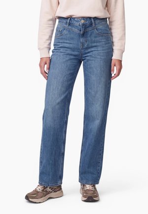 Person iført blå jeans med lige ben, lysebeige sweatshirt og brune sneakers, stående mod en ensfarvet hvid baggrund.