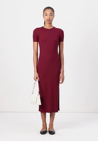 Burgundy ribbed midi dress with short sleeves, side slits, and a round neckline; paired with a small white handbag and black flats.