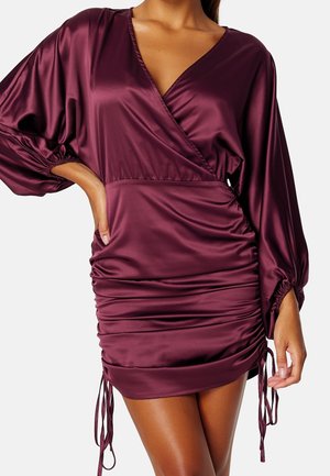 Femme portant une robe portefeuille en satin violet profond avec de longues manches bouffantes et des côtés froncés noués avec des cordons.