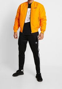 Orange bomberjacka med ribbade mudd, svart grafisk t-shirt, svarta byxor med vita ränder och svarta träningsskor.