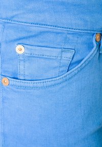 Jeans de mezclilla azul claro con un bolsillo frontal, costuras visibles y un herraje en tono dorado contrastante en el bolsillo. Textura suave.